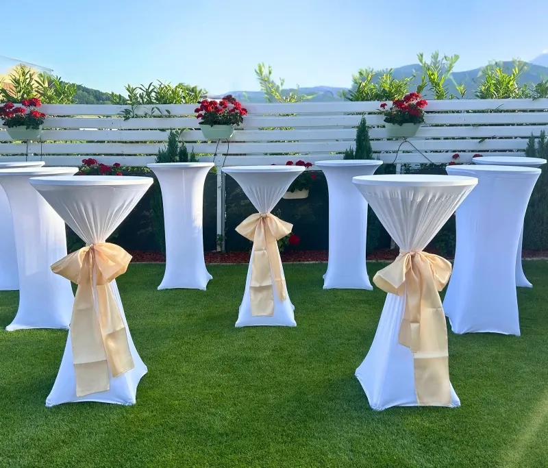 Décoration mariage : tables hautes à housse blanche ornées de nœuds dorés sur une pelouse décorée de fleurs