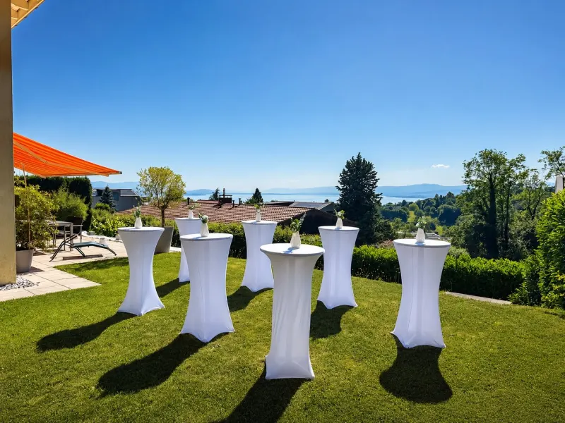 Anniversaire : tables hautes à housse blanche sur la pelouse d'un jardin avec vue sur le lac