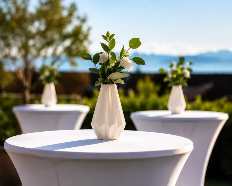 Anniversaire : tables hautes à housse blanche décorées de vases et de fleurs blanches, en plein air avec vue sur le lac